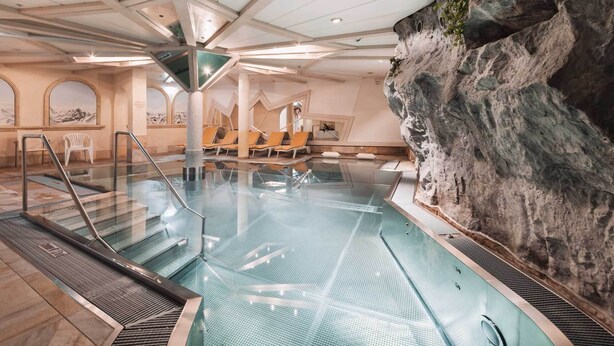 An indoor swimming pool with a rocky wall, lounge chairs, and a mezzanine level with seating. - Hotel Piz Buin (Ischgl)
