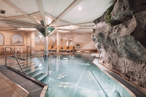 An indoor swimming pool with a rocky wall, lounge chairs, and a mezzanine level with seating. - Hotel Piz Buin (Ischgl)