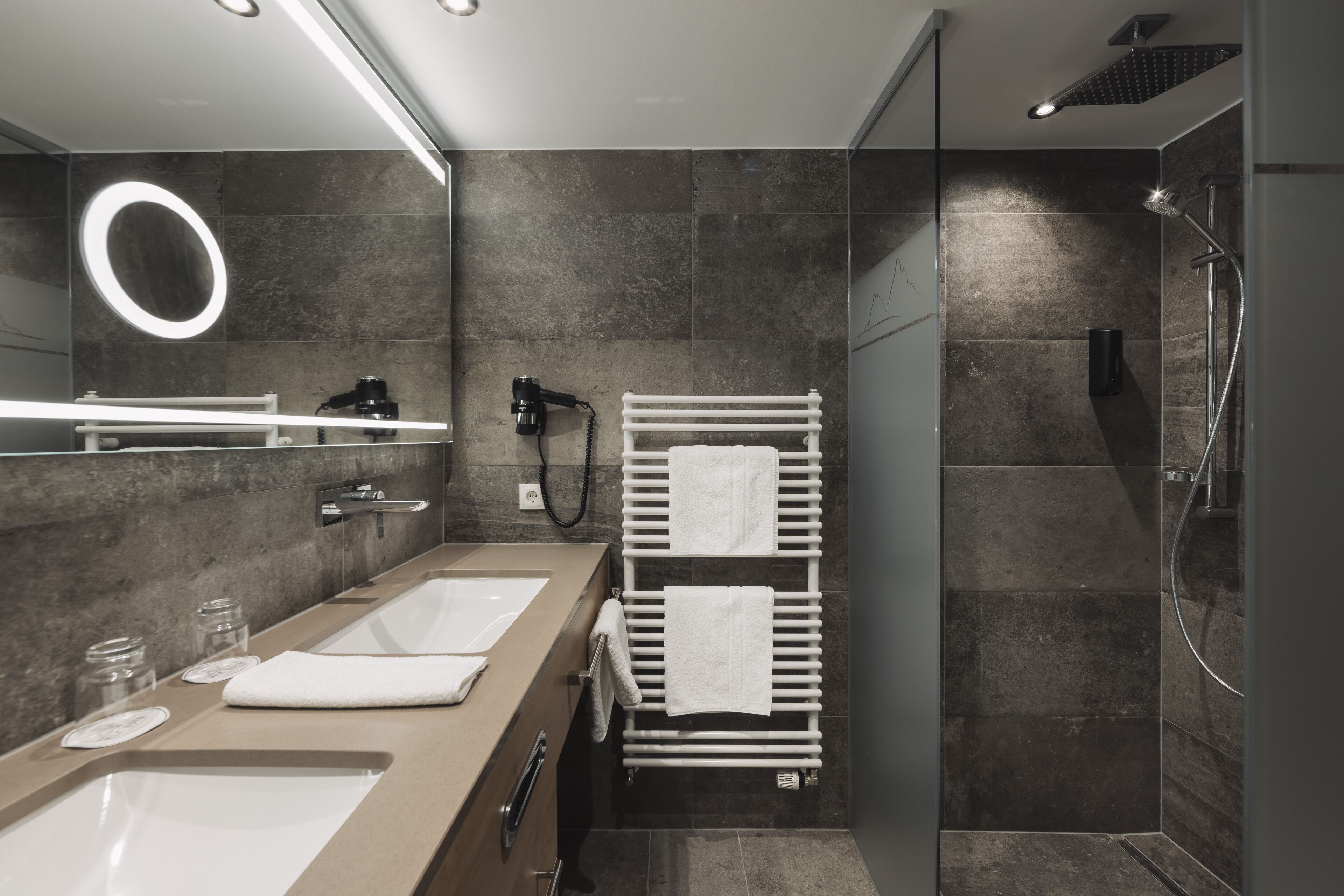 A modern bathroom with dual sinks, a large mirror, a towel rack, and a shower area.