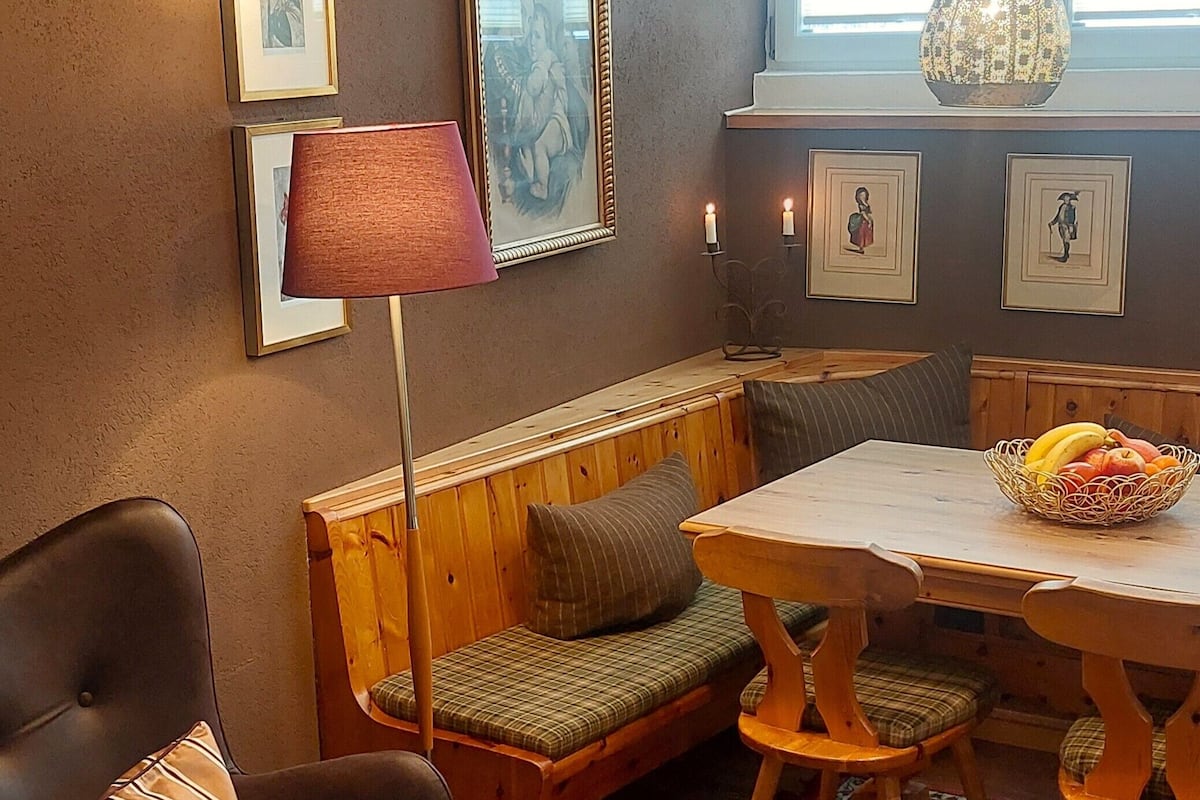 A cozy room with a wooden bench, a dining table with a fruit basket, a patterned rug, and framed artwork on the walls.