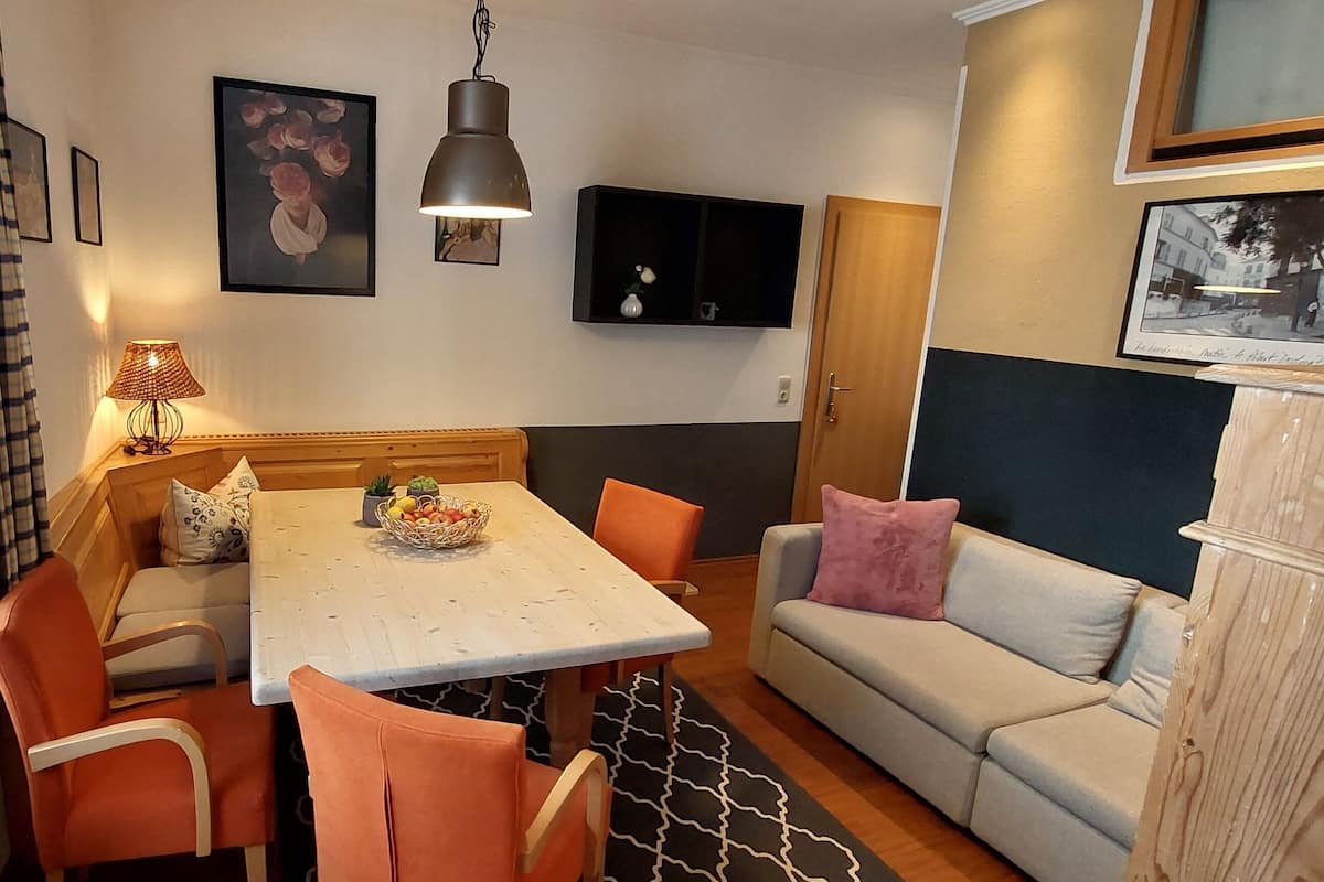 A cozy living room with a wooden dining table, orange chairs, a grey sofa, and a black and white patterned rug.