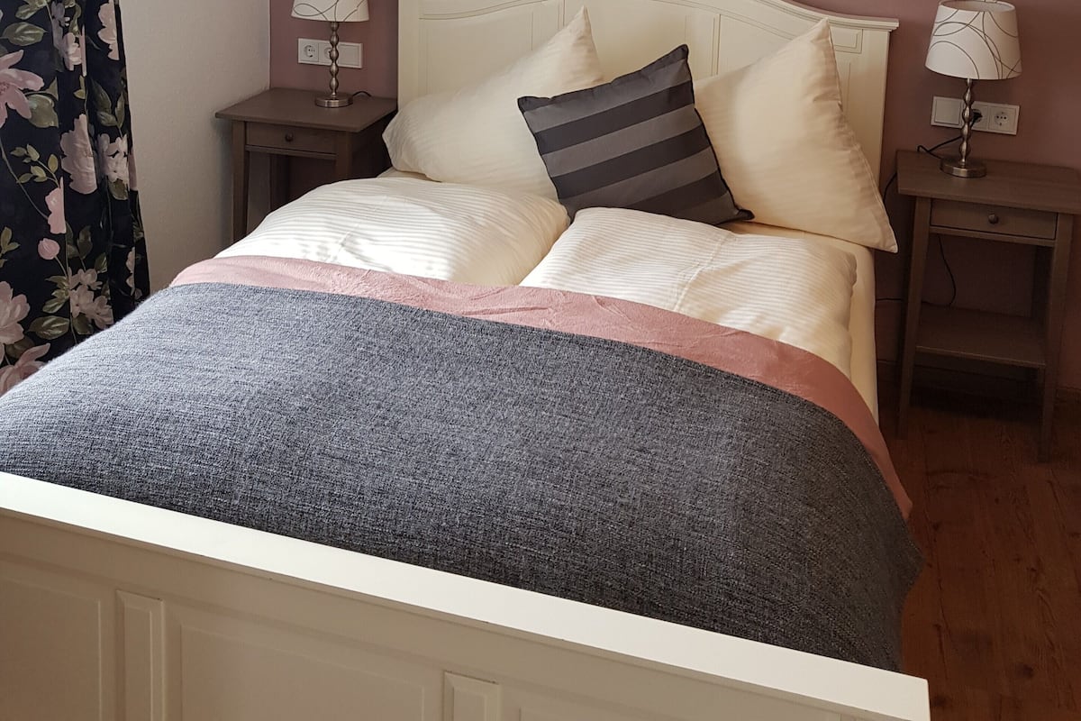 A bedroom with a bed, two bedside tables, a nightstand, a wall-mounted painting, and a floral curtain.