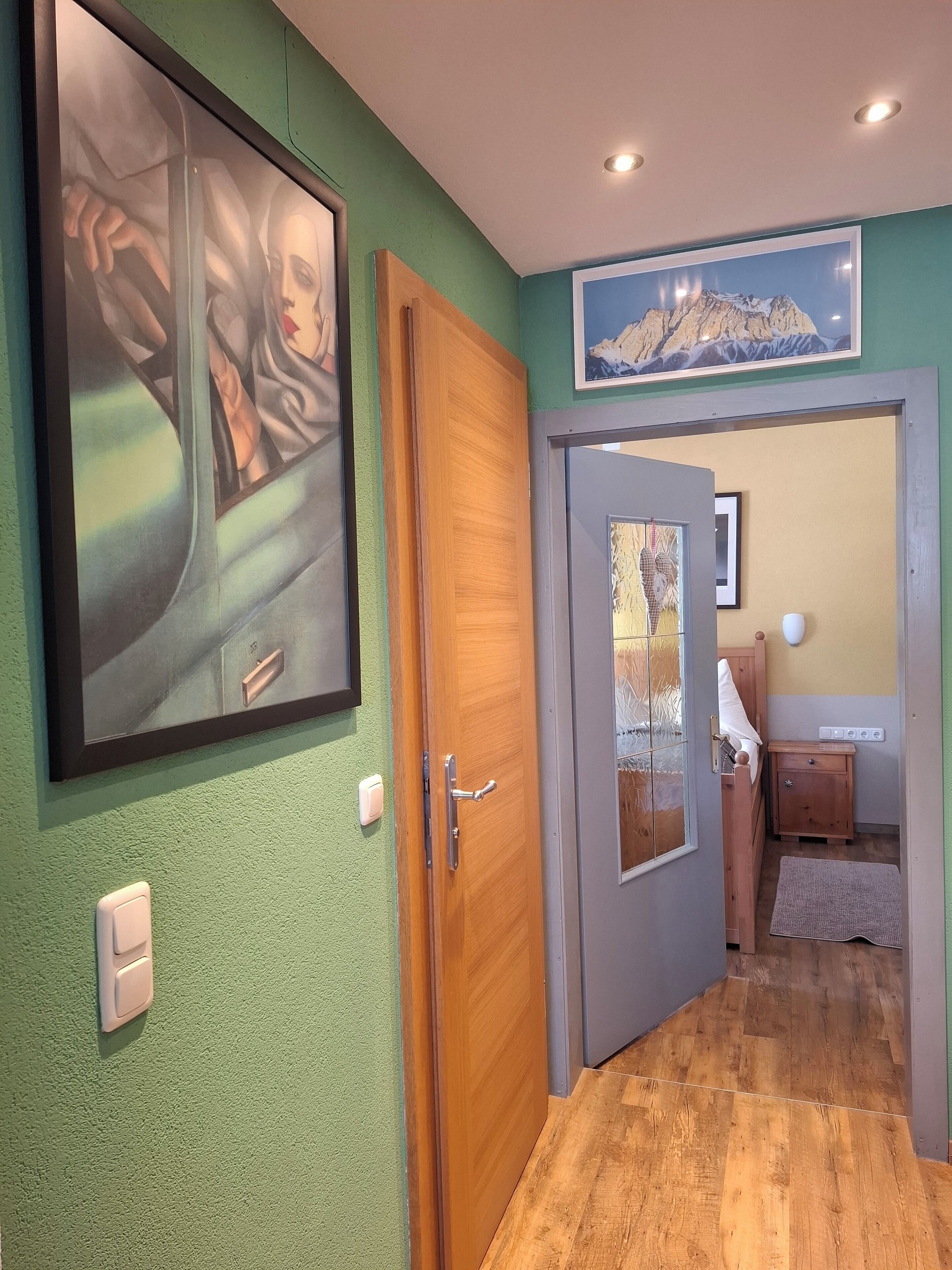 Un couloir avec un mur vert, une porte en bois et un tableau représentant une montagne.