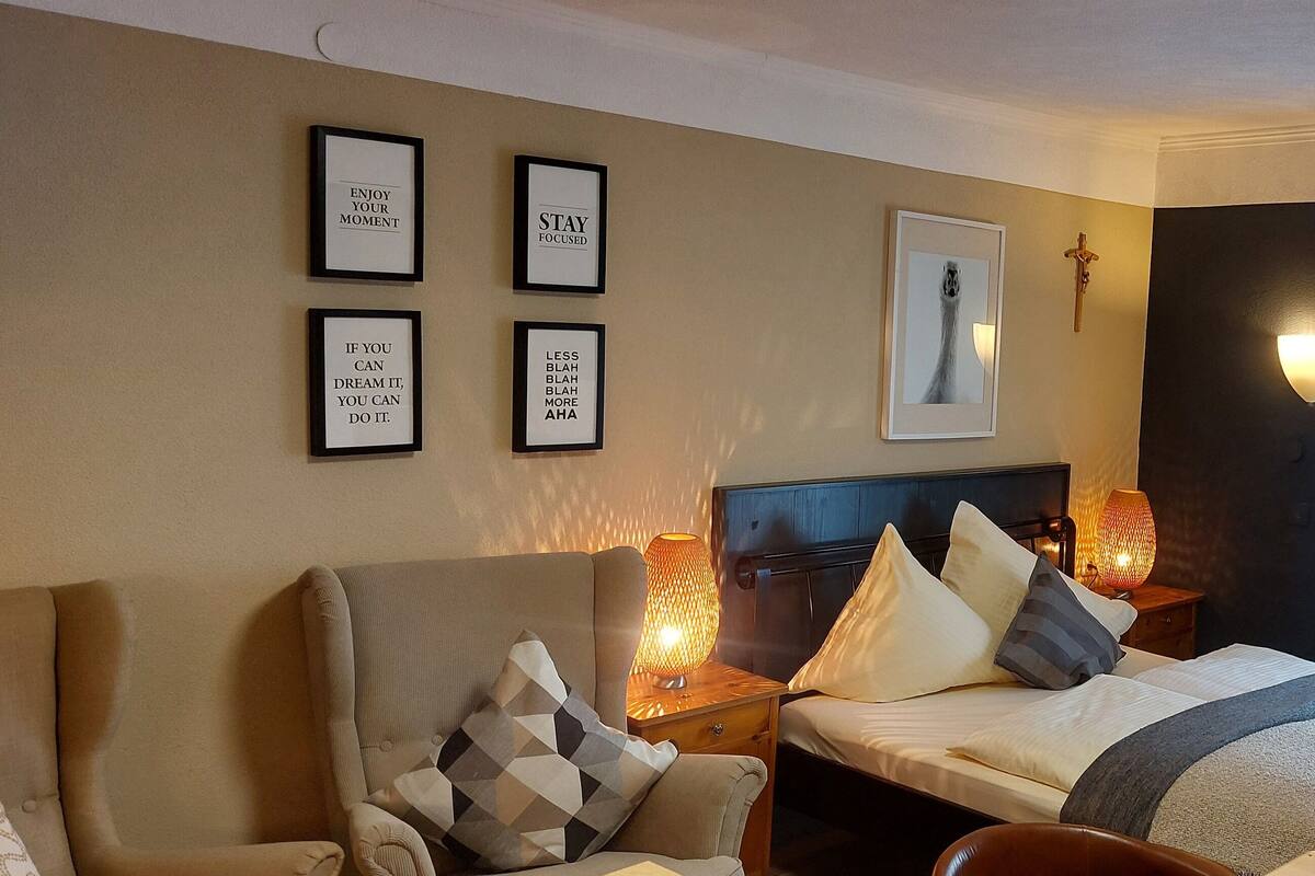 A hotel room with a bed, a sofa, and a nightstand with a lamp. There are framed motivational quotes on the wall.