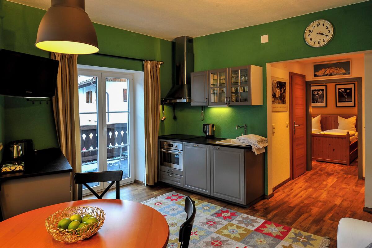 A compact kitchen with a dining area, a clock on the wall, and a view of the outside through a sliding door.