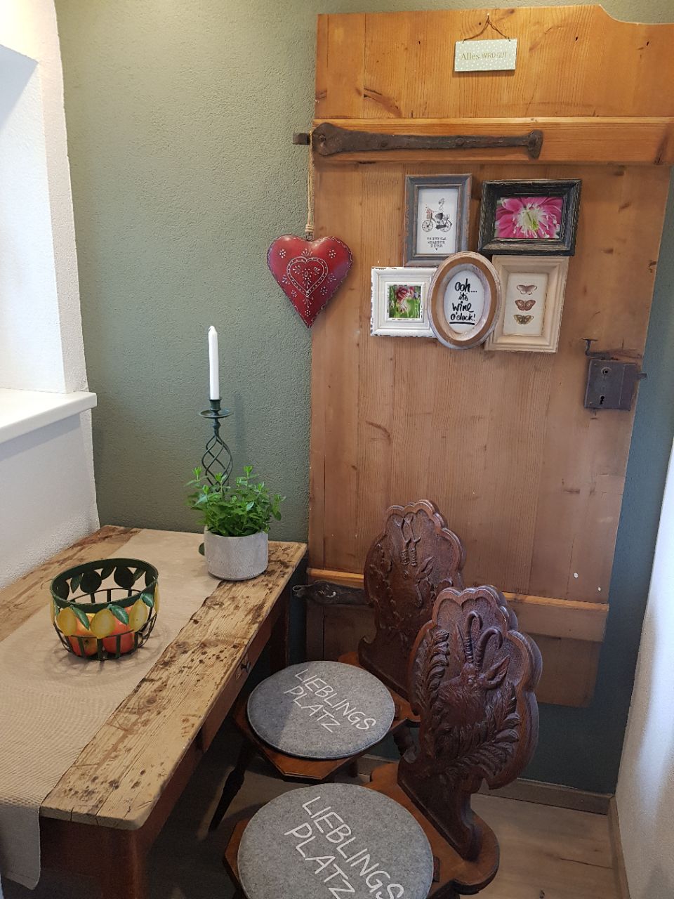 Rincón de una habitación con una puerta de madera, una mesita con una planta en maceta y dos sillas con el grabado 'LEBLINGS PLATZ'.