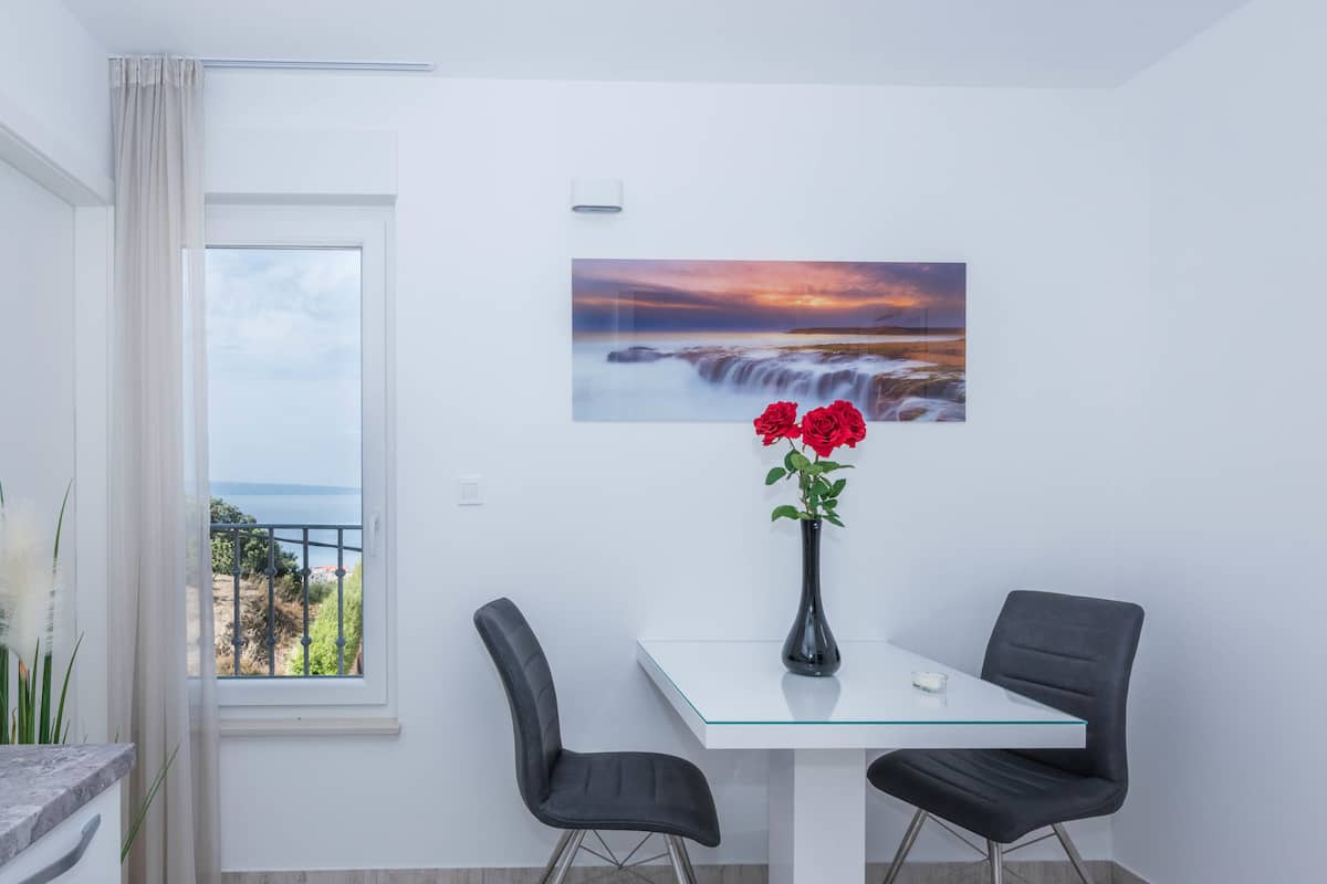 Un espace repas moderne avec une table en verre, deux chaises noires, un vase rempli de fleurs rouges et un tableau représentant un coucher de soleil accroché au mur.