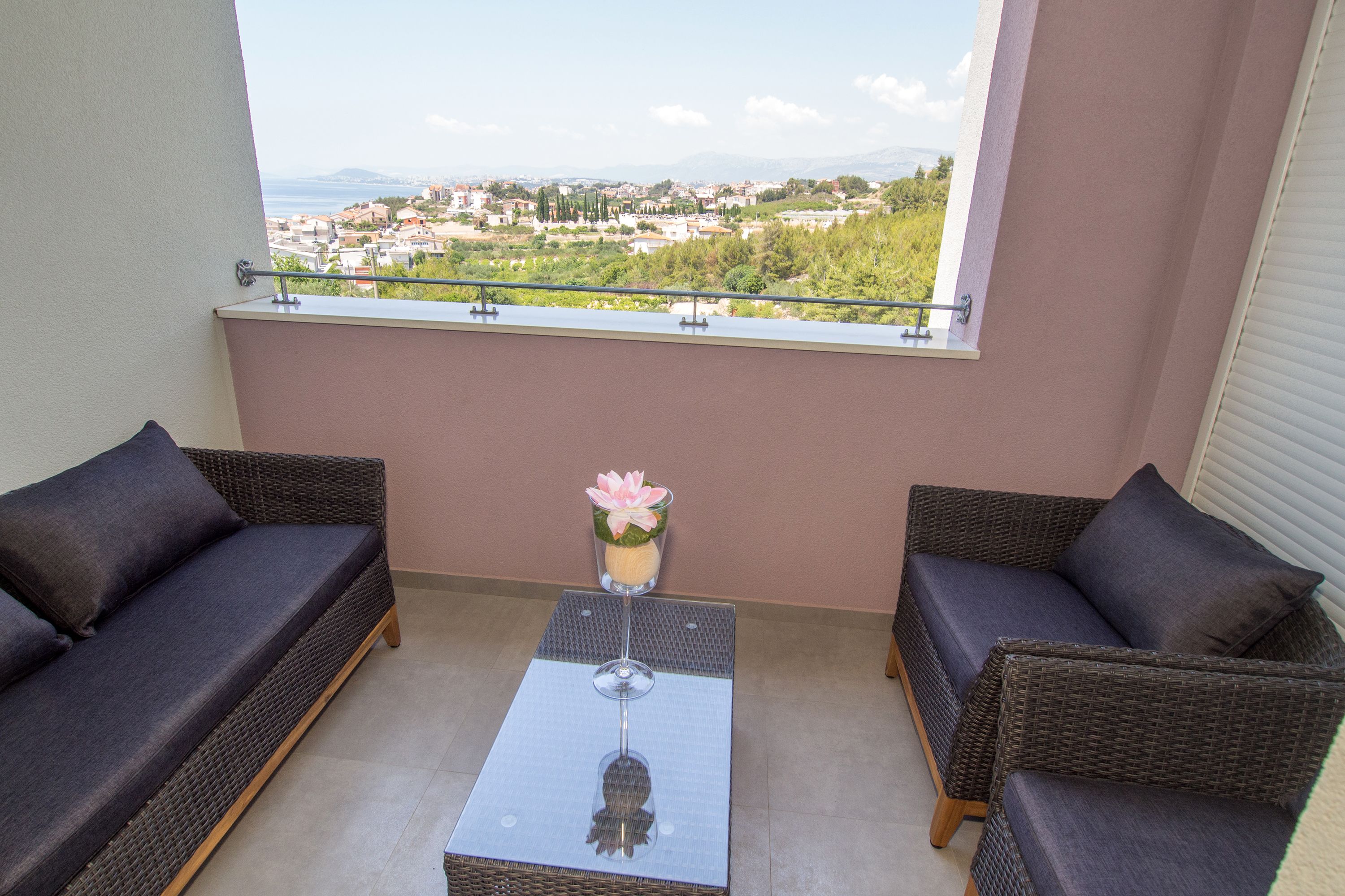 Een balkon met rieten meubels, een glazen tafel en uitzicht op een stad en bergen.