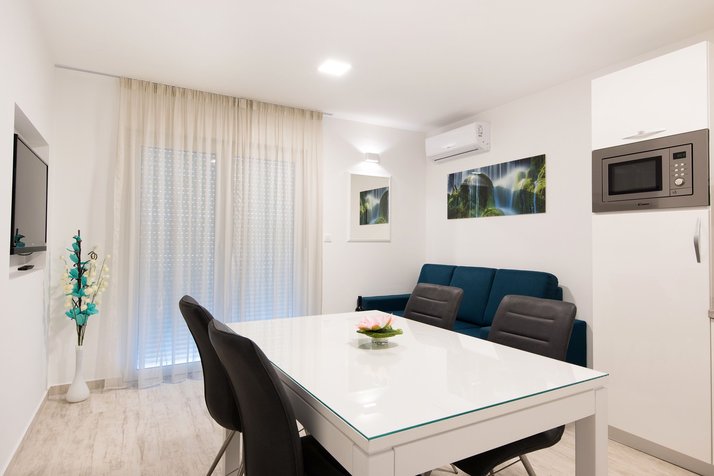 Ein moderner, gut beleuchteter Raum mit Esstisch, Stühlen, einem blauen Sofa und einem Fernseher.