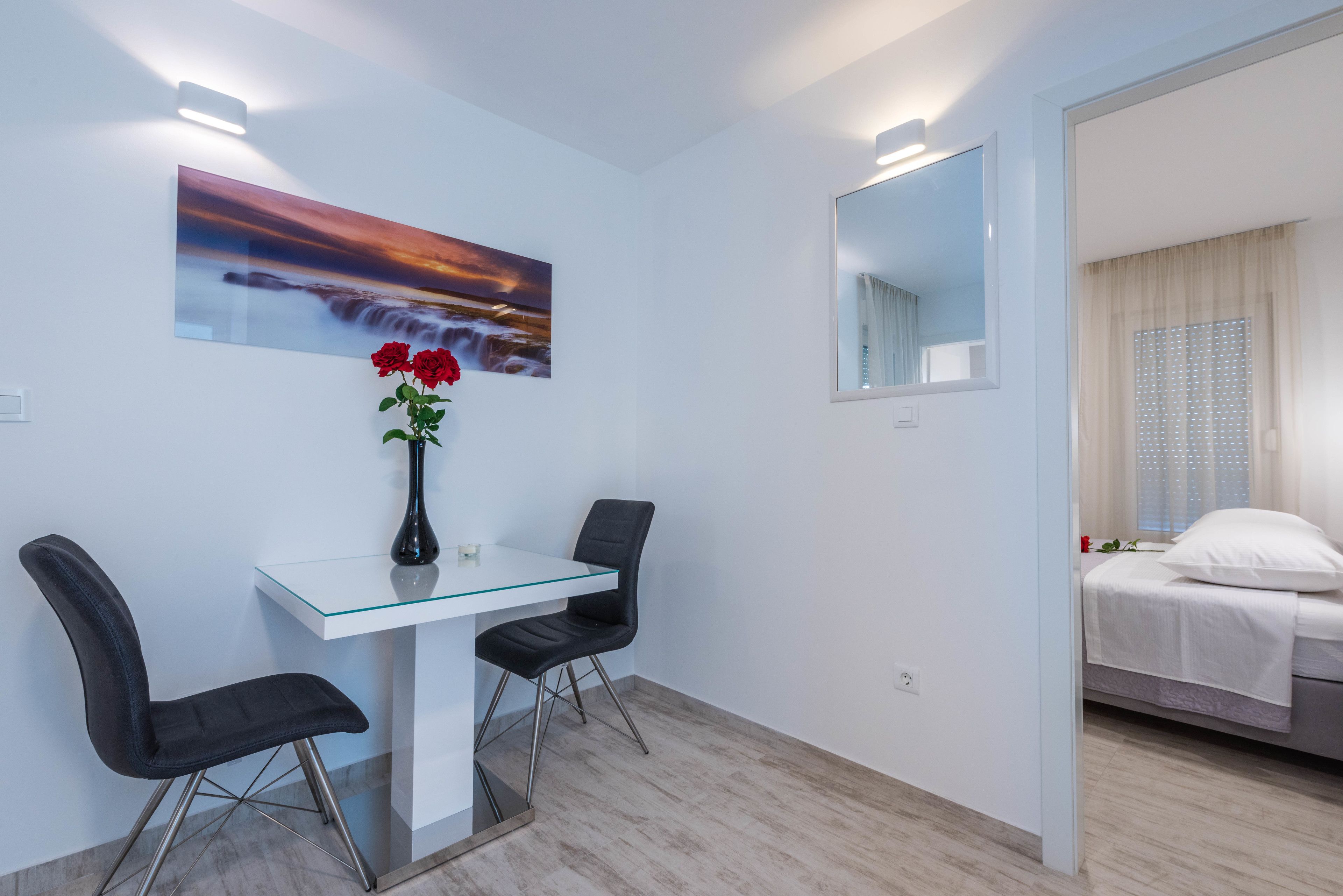 Et moderne hotelværelse med et sofabord i glas, to stole, en vase med røde blomster, et maleri af en solnedgang, et stort spejl og en seng med hvide sengetøj.