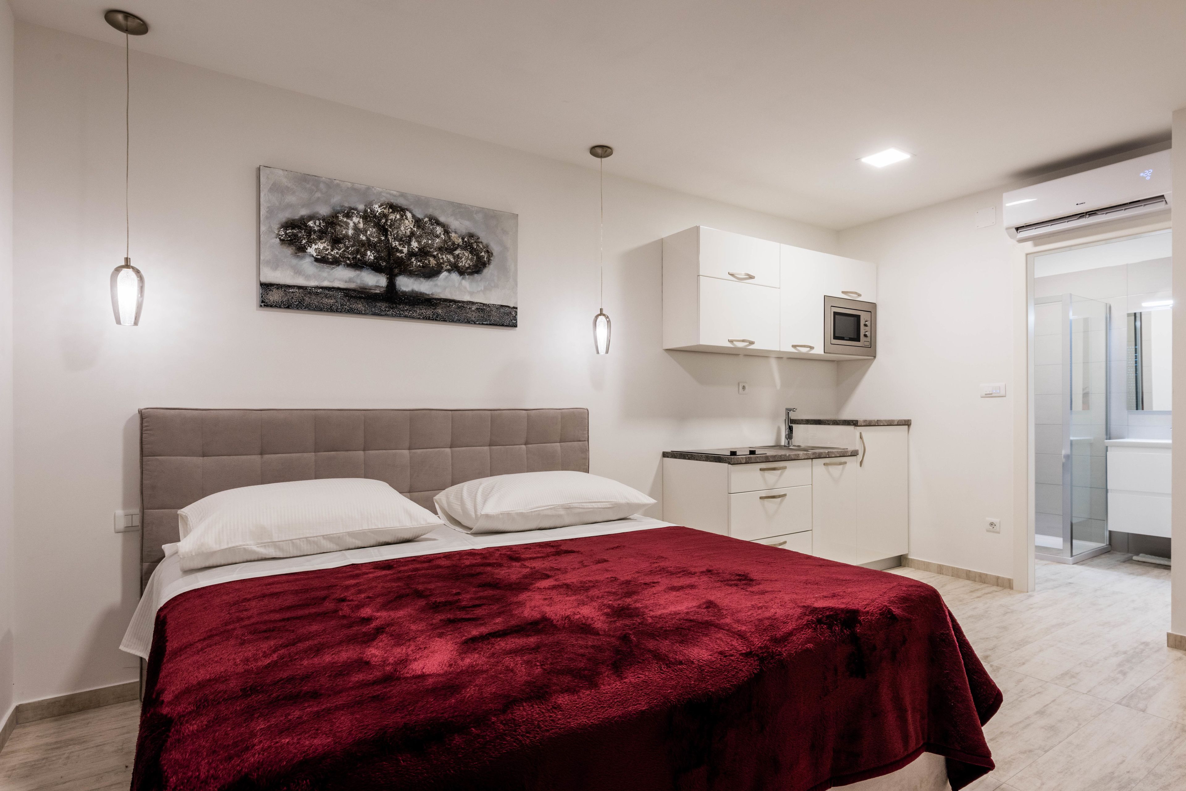 Habitació amb llit, manta vermella, un quadre, armari blanc, microones i bany amb dutxa de vidre.