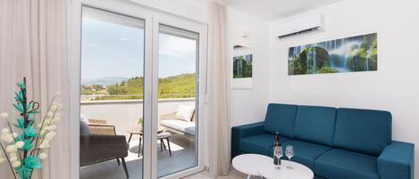 A modern living room with a blue sofa, a small round table with two wine glasses, and a view of a balcony with outdoor furniture.