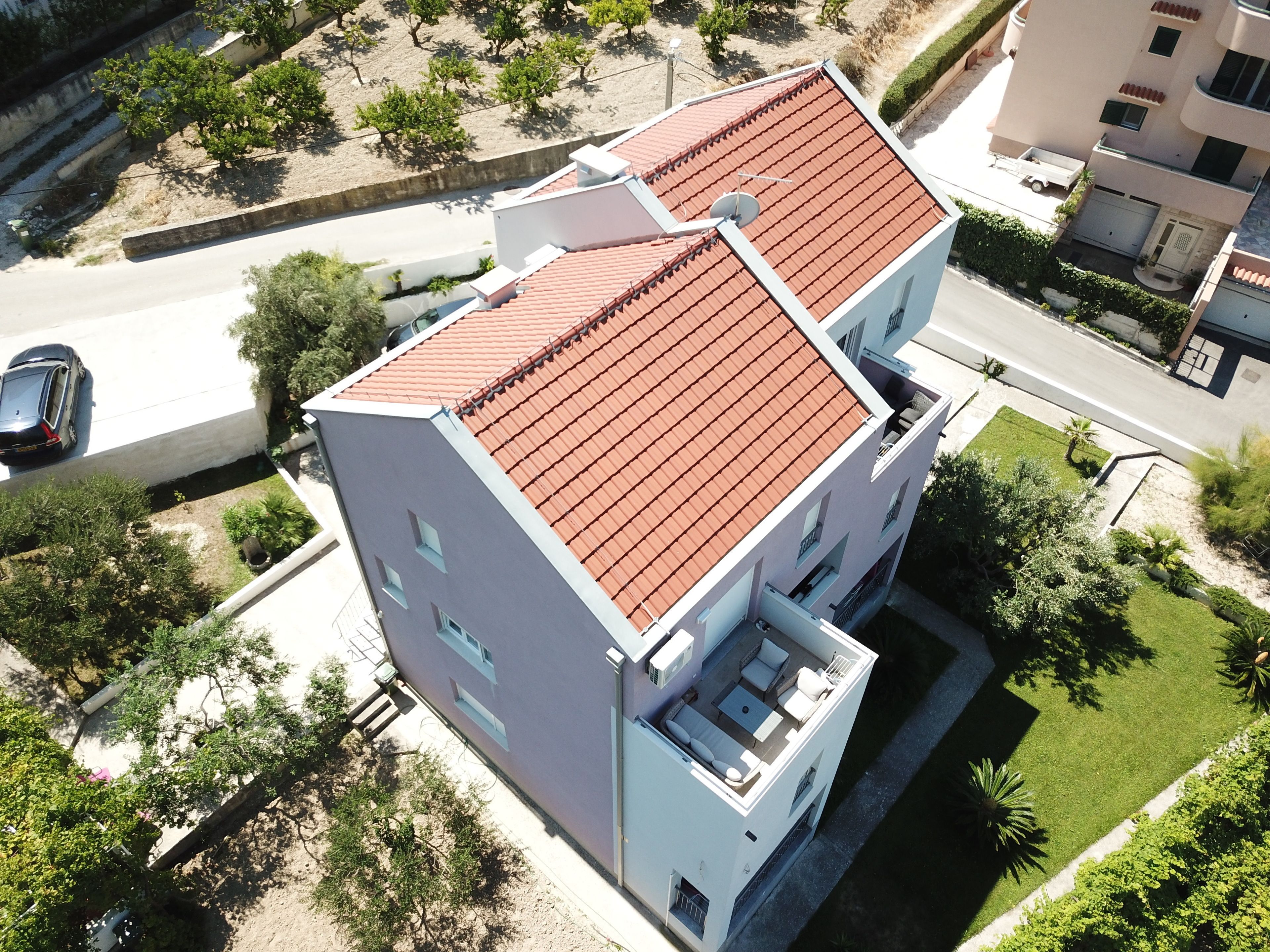 A modern two-story building with a red-tiled roof, a flat roof terrace with outdoor furniture, and a well-maintained lawn.