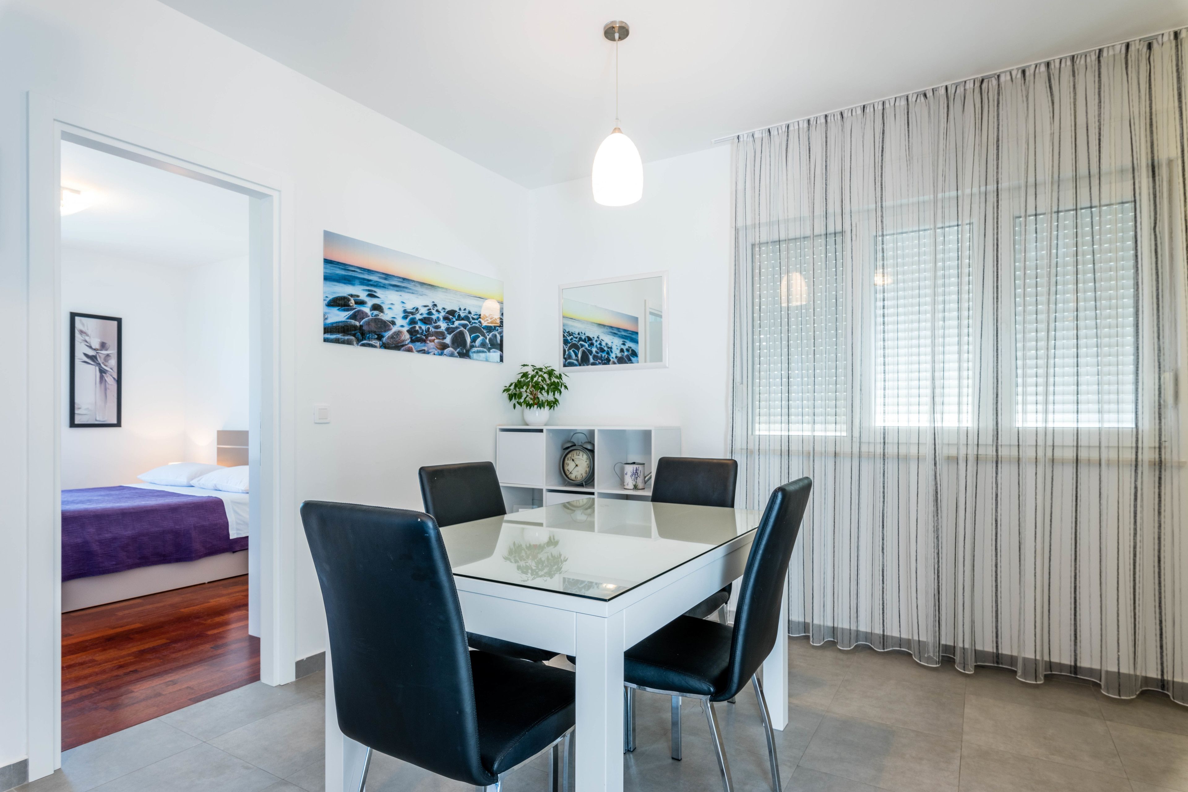 A modern dining area with a glass table and black chairs, a bed with a purple blanket in the adjacent room, and a large window with sheer curtains.