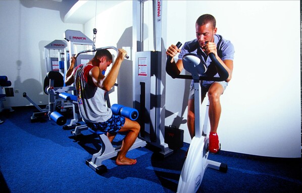 Two individuals exercising on stationary bikes in a gym with various equipment.