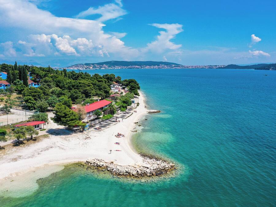 A coastal area with a beach, clear blue water, and buildings with red roofs.