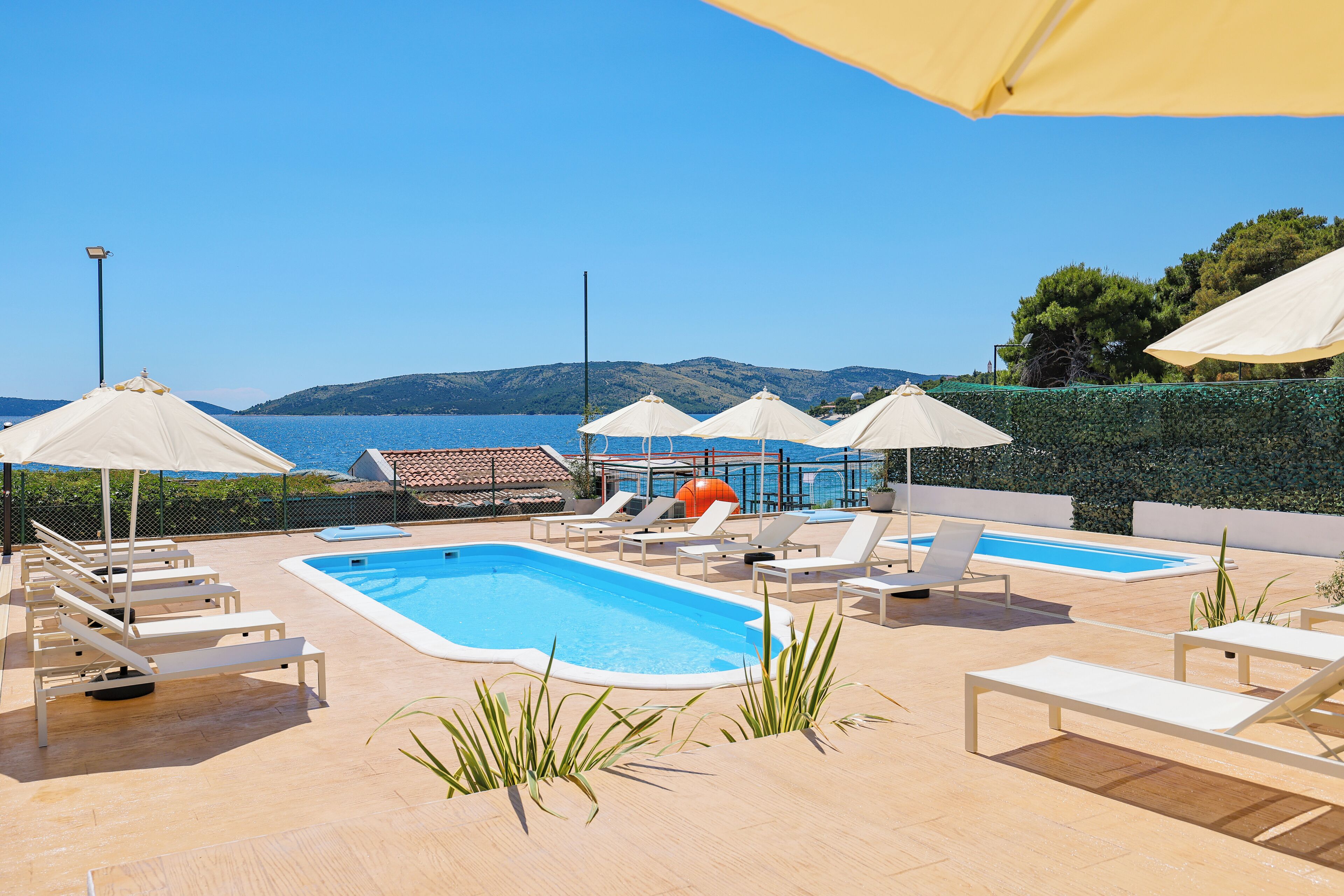 A poolside area with lounge chairs and umbrellas, a small pool, and a view of the sea and hills in the background.