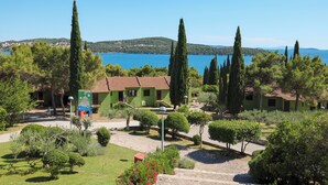 A courtyard with green buildings, a clear blue sky, and a lake in the background. - Apartments Medena (Seget)