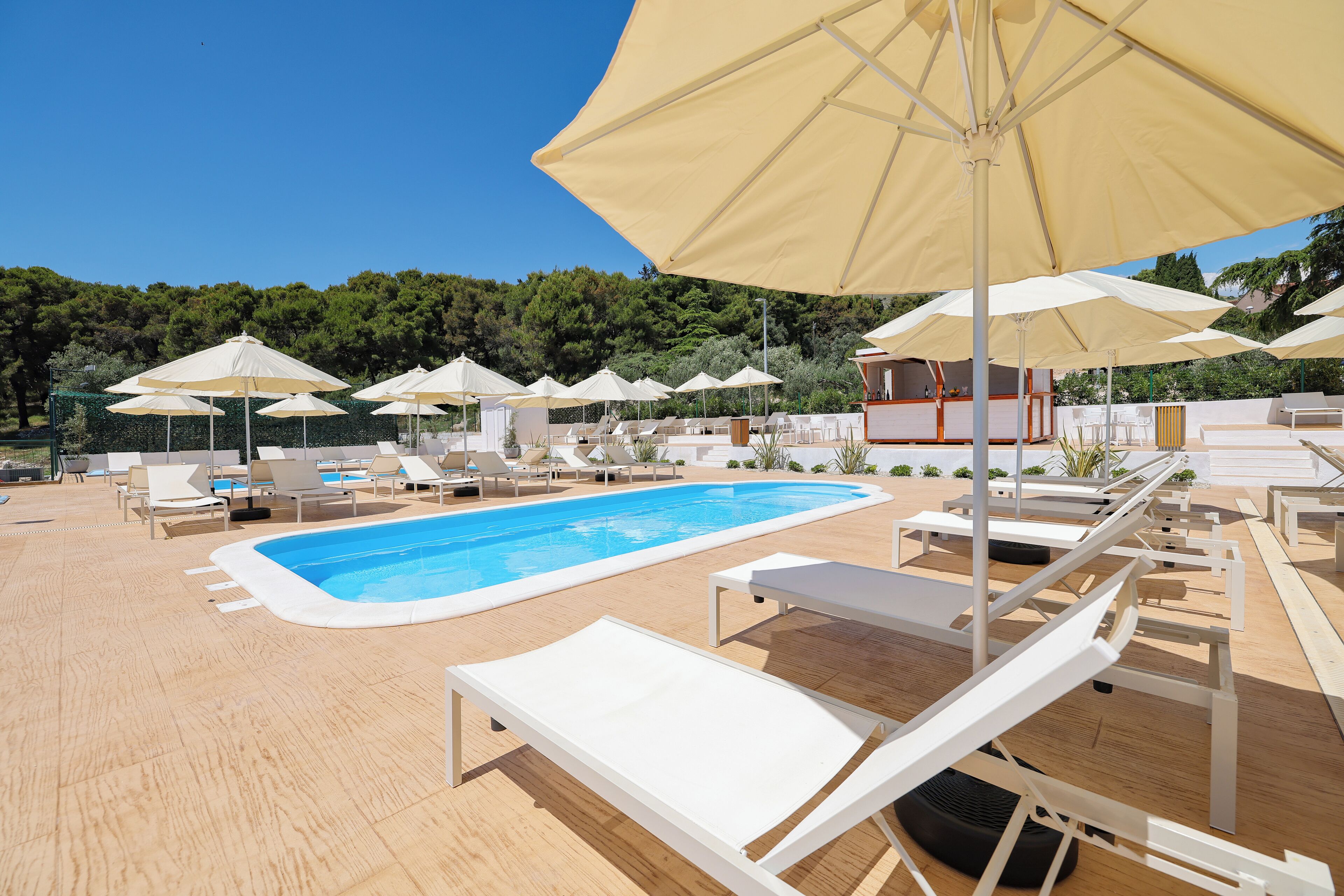 Zona de piscina con tumbonas y sombrillas, rodeada por una terraza de madera y una piscina.