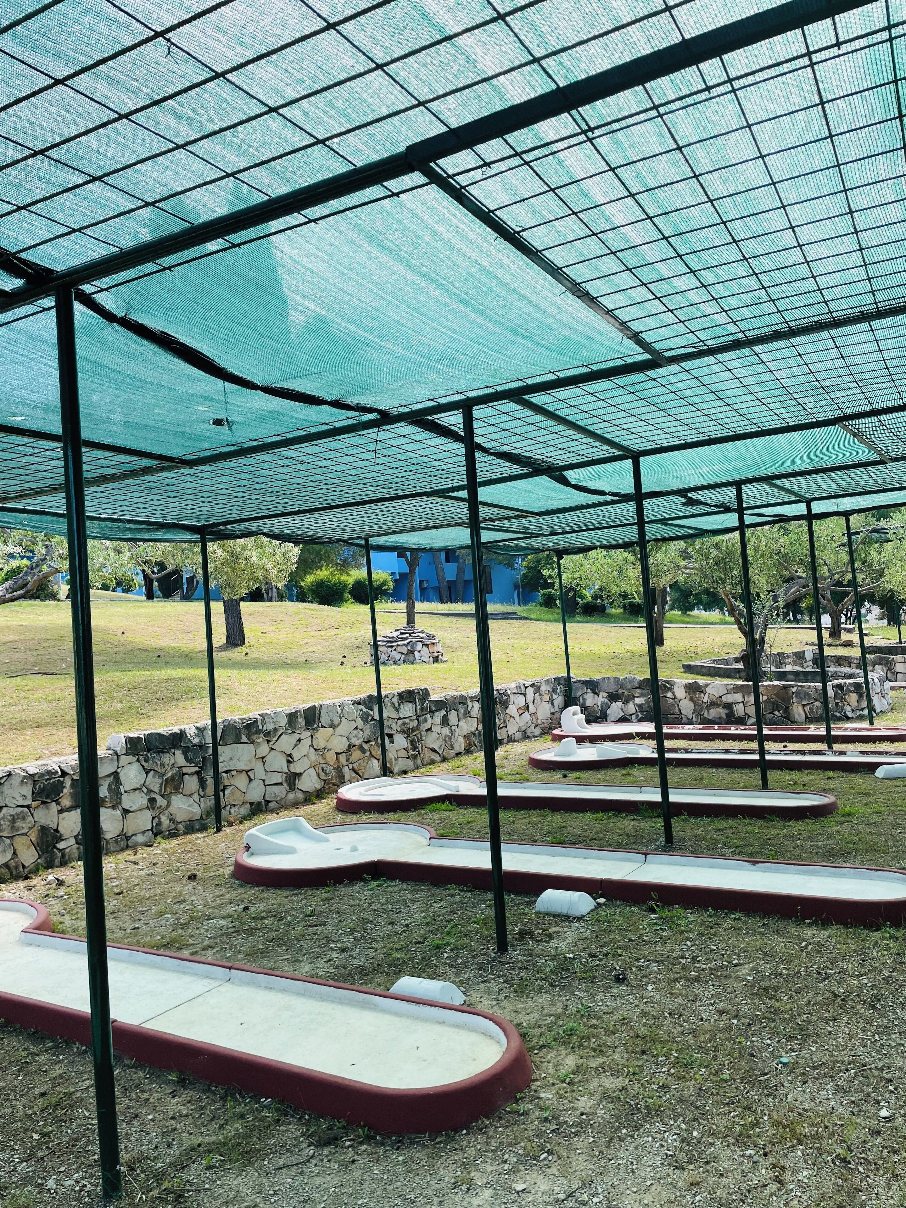 A covered outdoor area with a grid-like ceiling structure, stone walls, and a grassy field in the background.