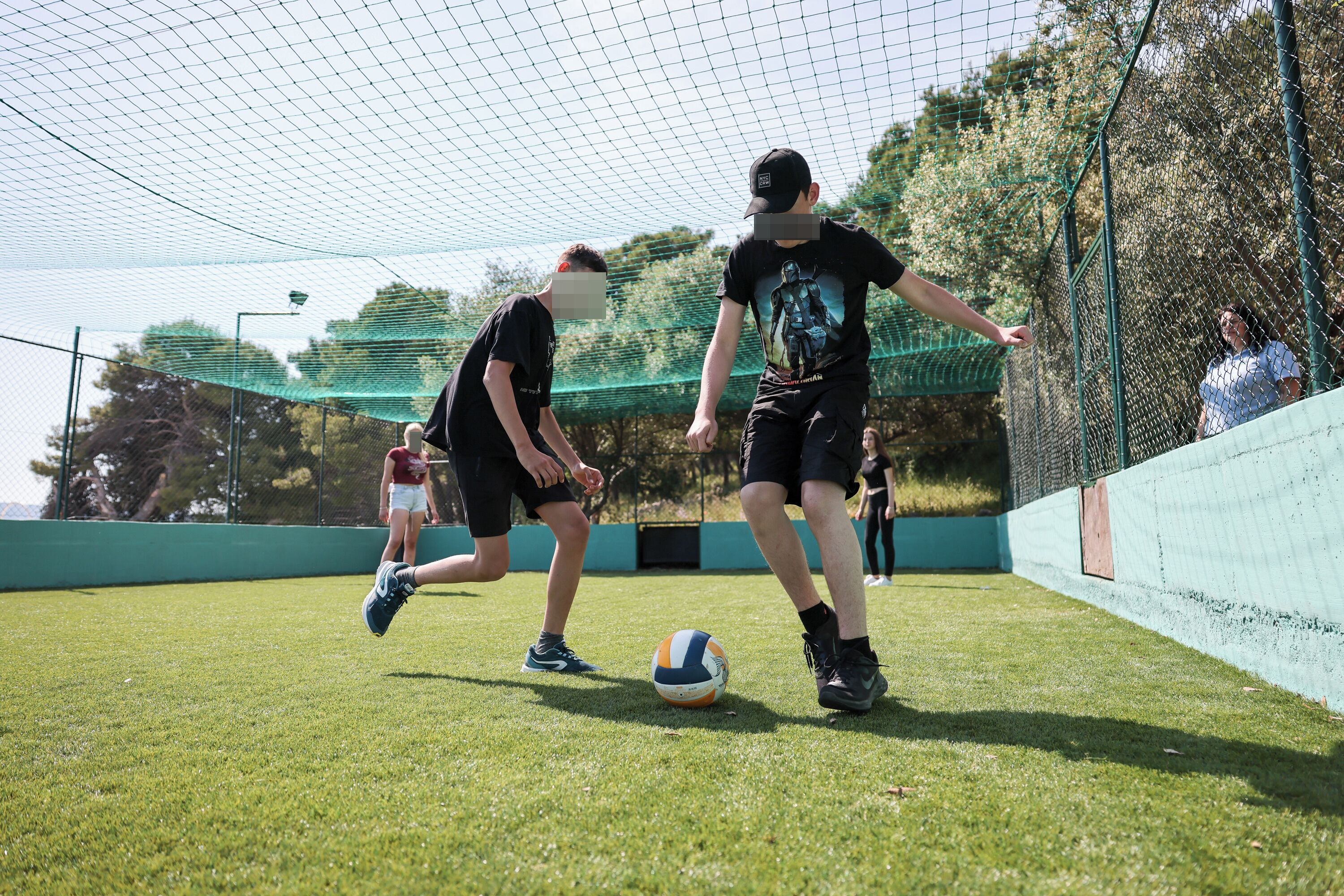 A netted sports area with a volleyball on the ground, surrounded by a green fence and trees in the background.