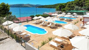 A pool area with multiple pools and lounge chairs, surrounded by umbrellas, with a view of the sea and hills in the background. - Apartments Medena (Seget)