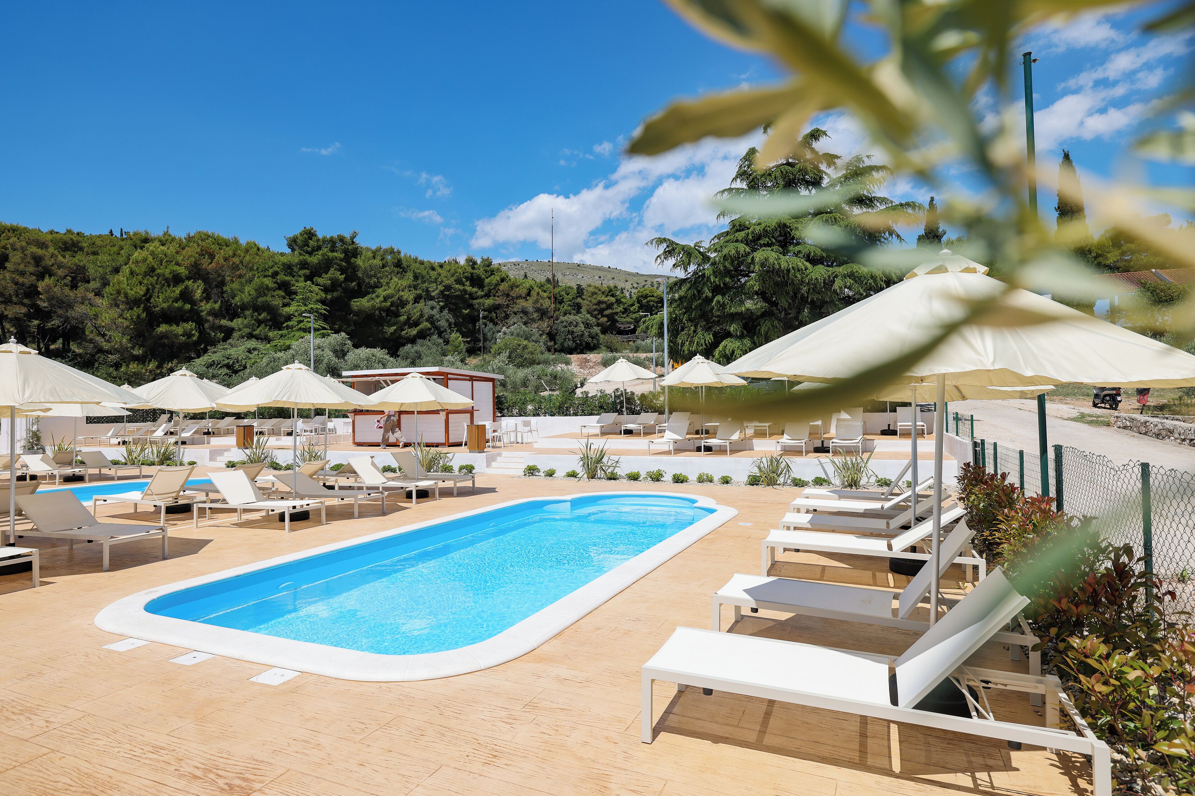 Un área de piscina con sillas de descanso y sombrillas, rodeada de follaje y un cielo despejado.