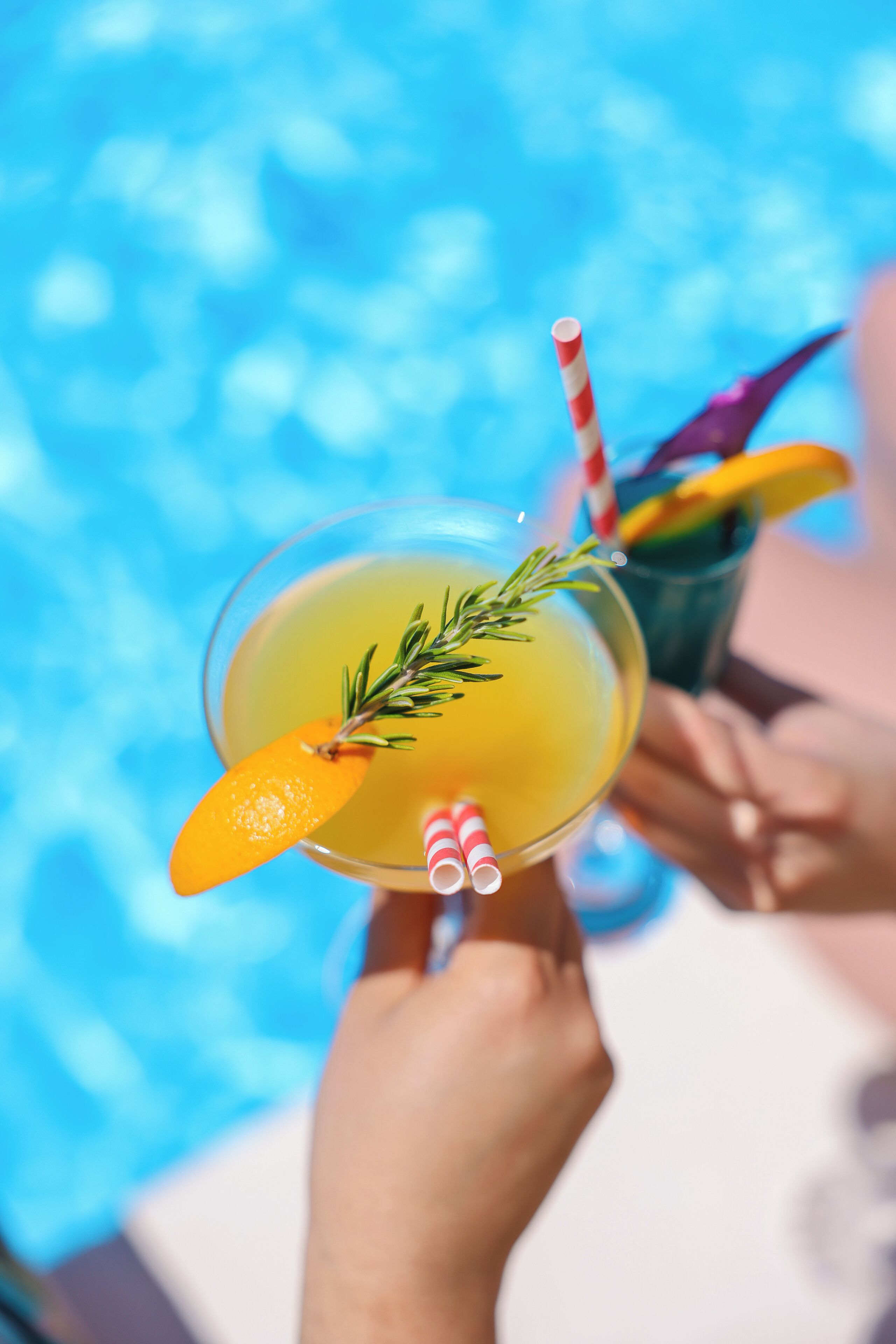 A cocktail with a slice of orange and a sprig of rosemary, held by a person with a striped straw.