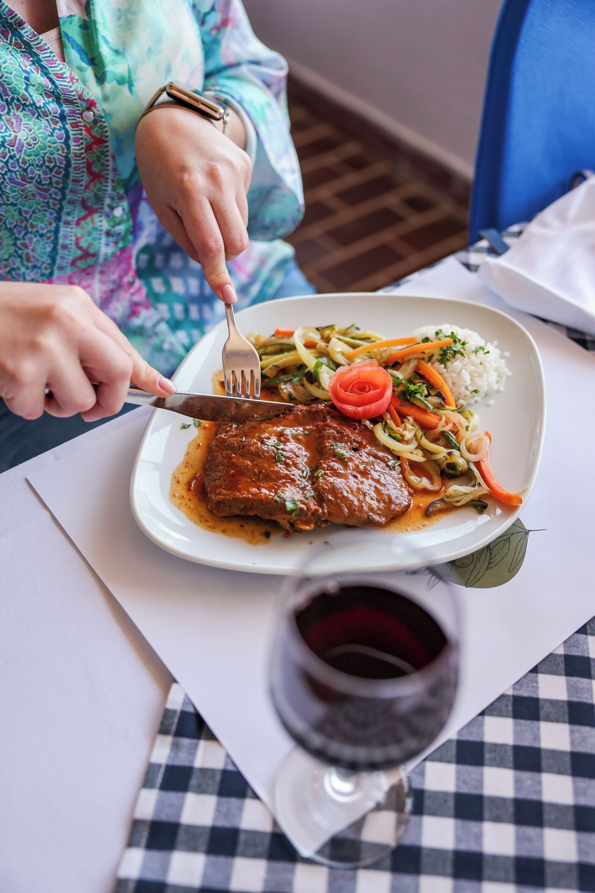 A person dining at a table with a plate of food, a fork, and a glass of red wine.