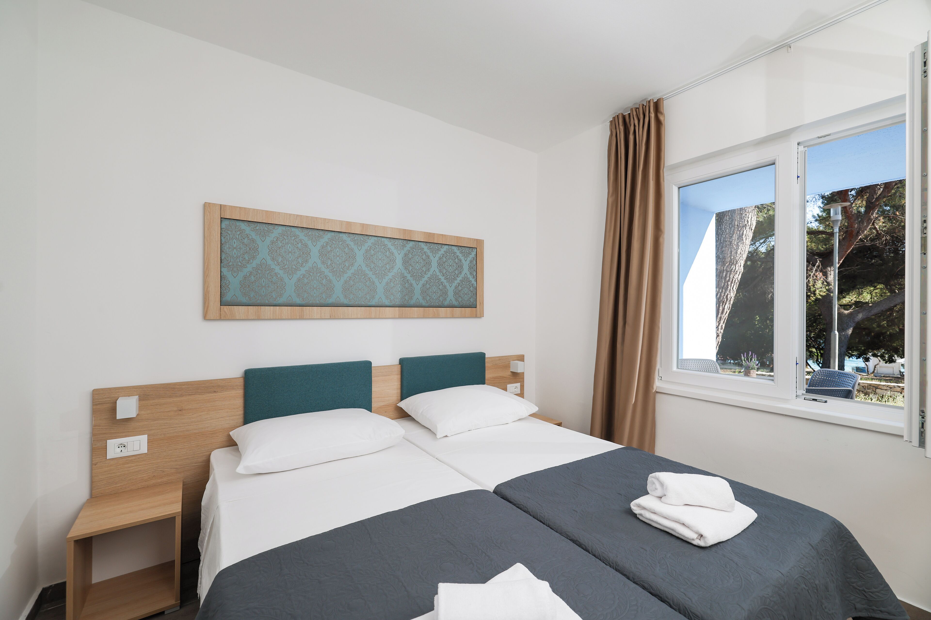 A double bed with two pillows, a nightstand, a wall-mounted light switch, a window with curtains, and a view of trees outside.