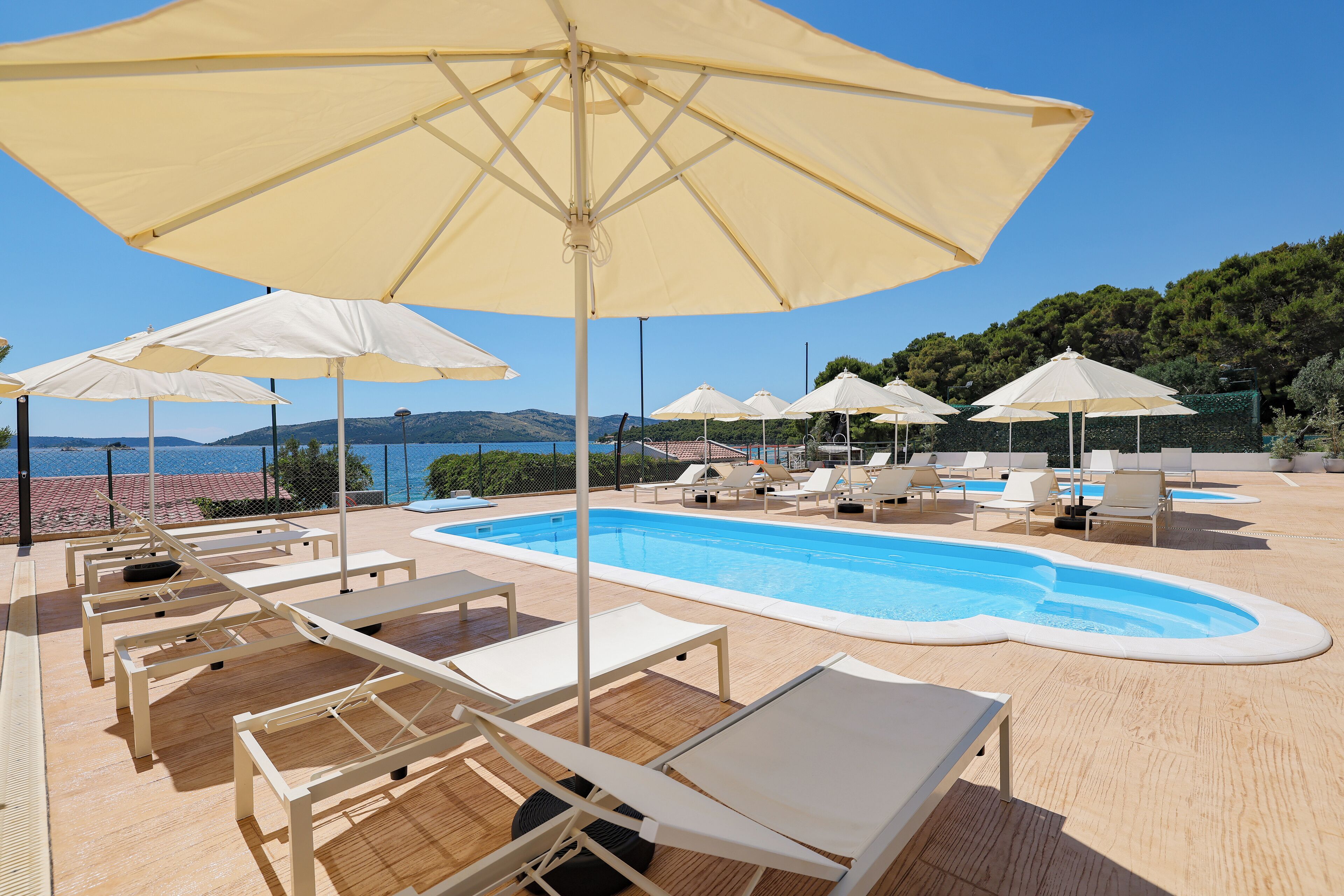 A poolside area with sun loungers and umbrellas, a swimming pool, and a view of the sea and hills.