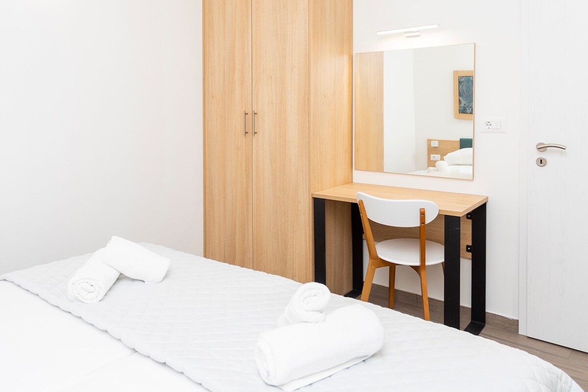 A hotel room with a bed, a desk, a chair, a wooden wardrobe, a mirror, and a door.