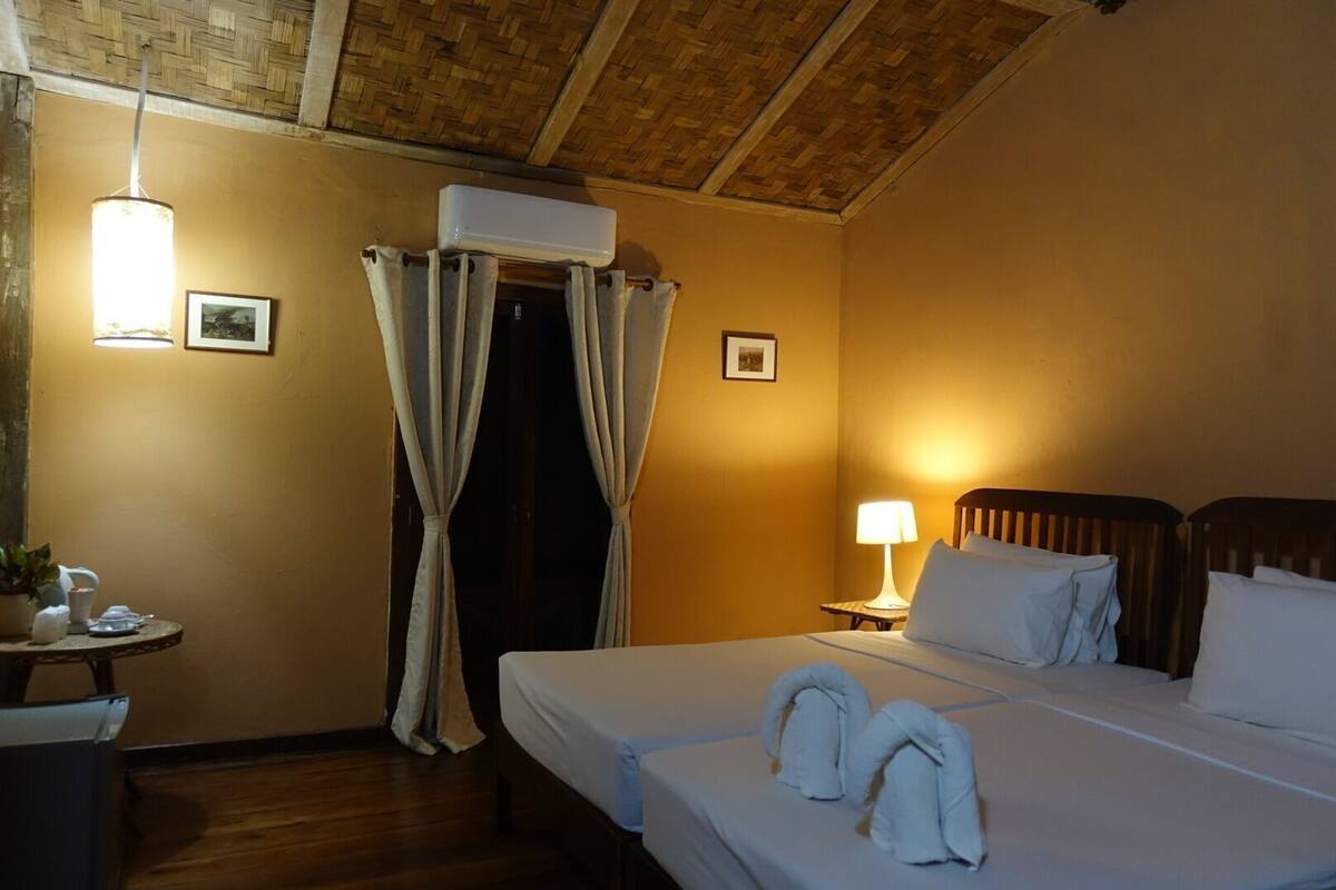 Habitación de hotel con cama, mesita de noche con lámpara, pequeña mesa con planta y ventana con cortinas.