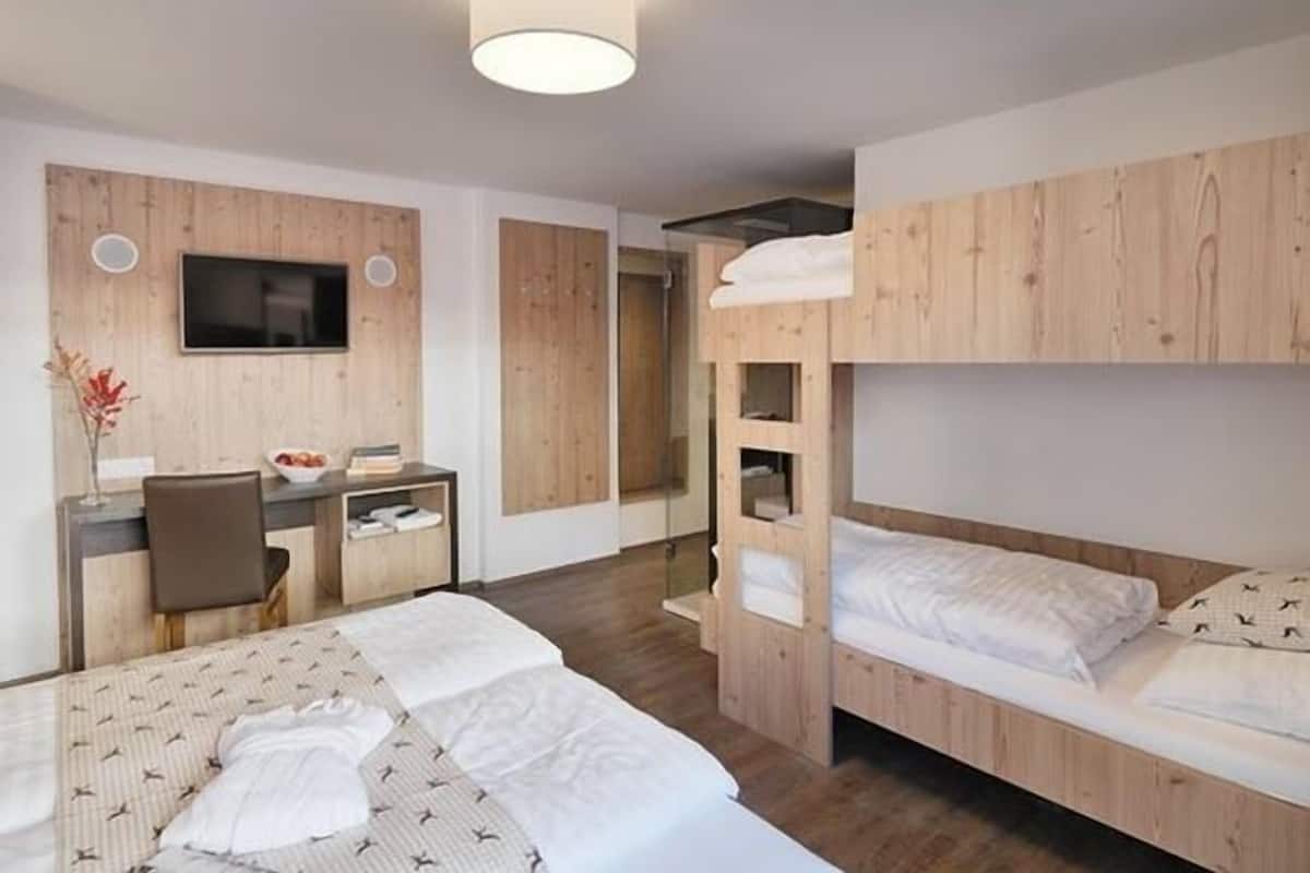 A modern hotel room with a bunk bed, a desk with a chair, a flat-screen TV mounted on the wall, and a bedside table with a vase of flowers.