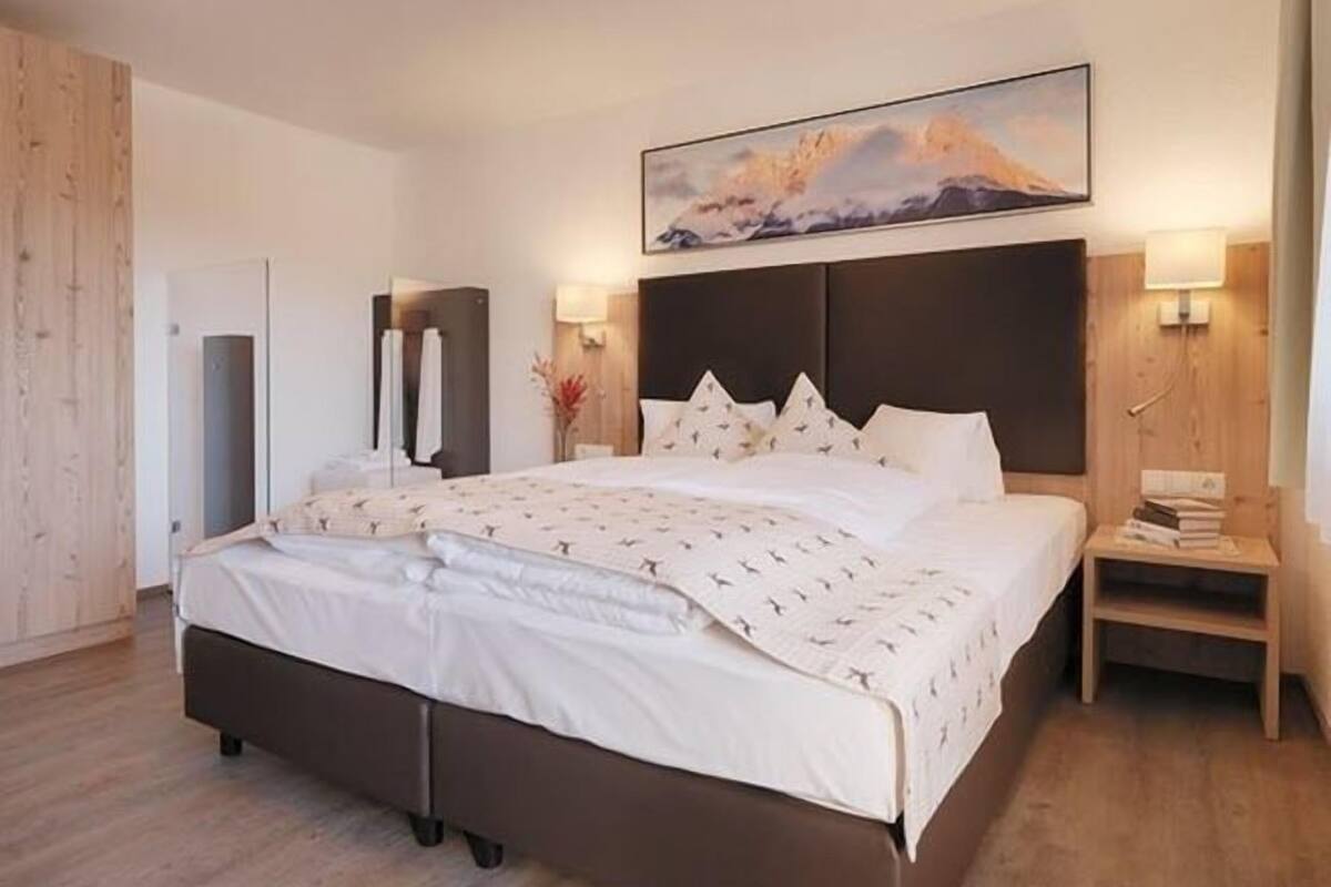 A modern hotel room with a large bed, a wooden headboard, a bedside table with a lamp, and a painting above the bed.