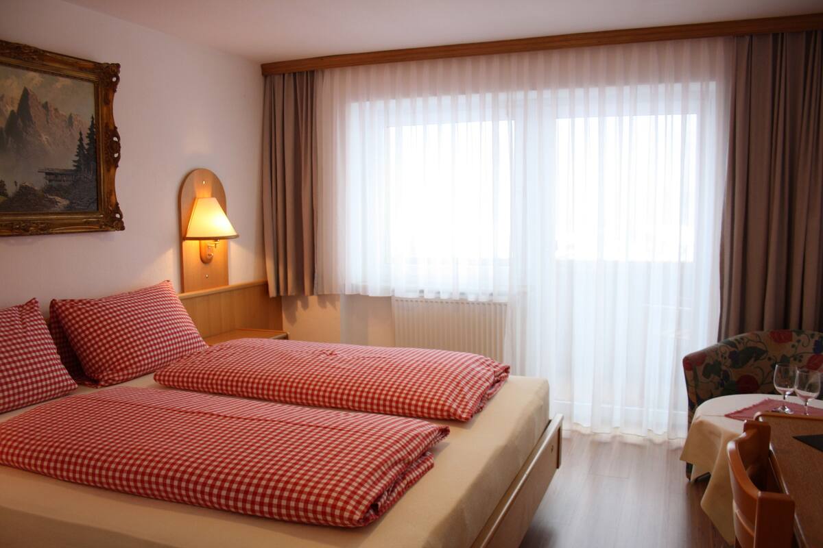 Habitación de hotel con dos camas, un cuadro en la pared, una lámpara, una ventana con cortinas y una mesita con una silla.