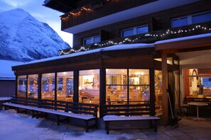 A snow-covered mountain at dusk, a wooden building with large windows, and benches outside. - Landhotel Wolf (Leutasch)