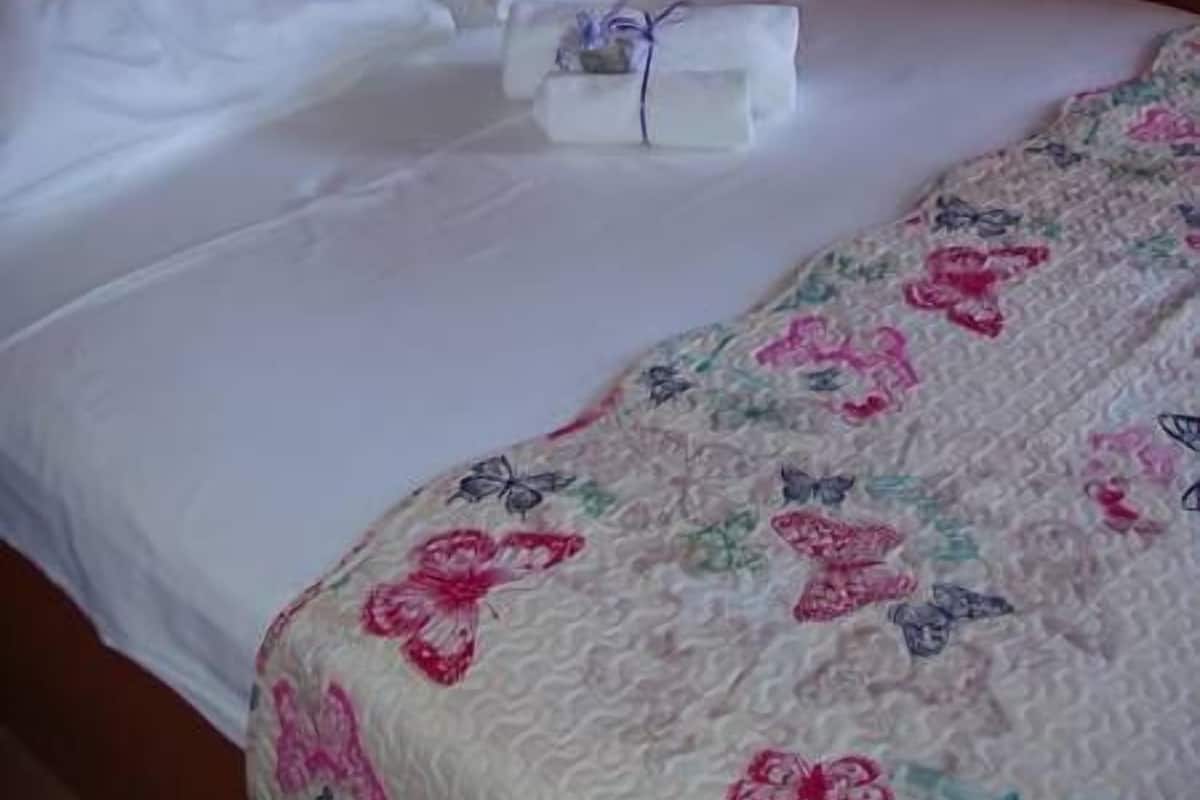 Un lit bien fait, avec des draps blancs et une couverture à motifs floraux. Un petit paquet blanc est posé sur le lit.