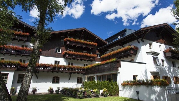 A multi-story building with balconies adorned with flowers, surrounded by a well-maintained lawn and trees. - Valluga (Sankt Anton am Arlberg)