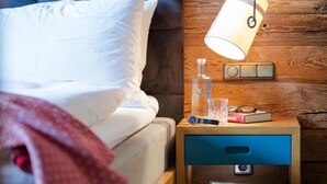 A neatly made bed with white linens, a bedside lamp, a bottle, a glass, and a book on a wooden nightstand with a blue drawer. - Valluga (Sankt Anton am Arlberg)