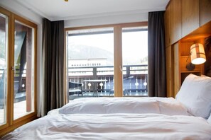 A hotel room with a bed, a window with curtains, a view of a building, and a balcony with a table and chairs. - Valluga (Sankt Anton am Arlberg)