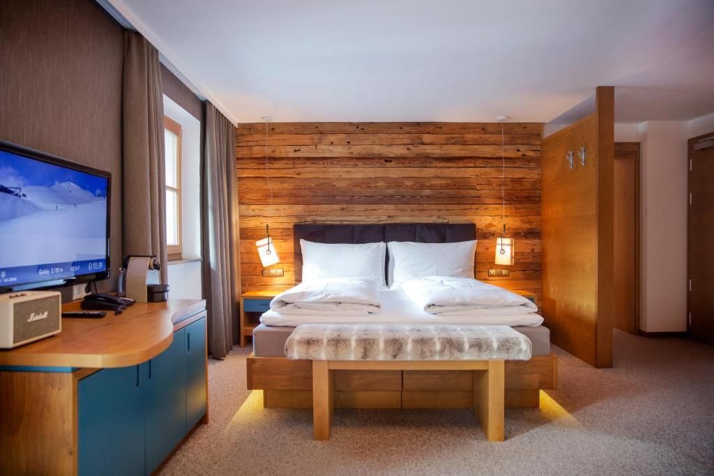 Una habitación de hotel con cabecera de madera, cama con ropa blanca, escritorio con televisor y mesita de noche con lámpara.