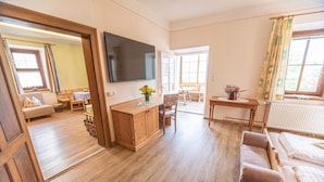 A room with a wooden floor, a television mounted on the wall, a wooden cabinet, a sofa, a chair, a table with a pitcher of flowers, and a window with curtains. - Hotel Gasthof Gribelehof (Lienz)