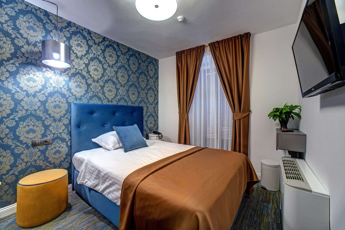 Une chambre d’hôtel avec un papier peint à motifs bleus, une tête de lit capitonnée, une couvre-lit marron, un petit pouf rond, une table de chevet avec une lampe, un téléviseur fixé au mur et une plante en pot posée sur une étagère.