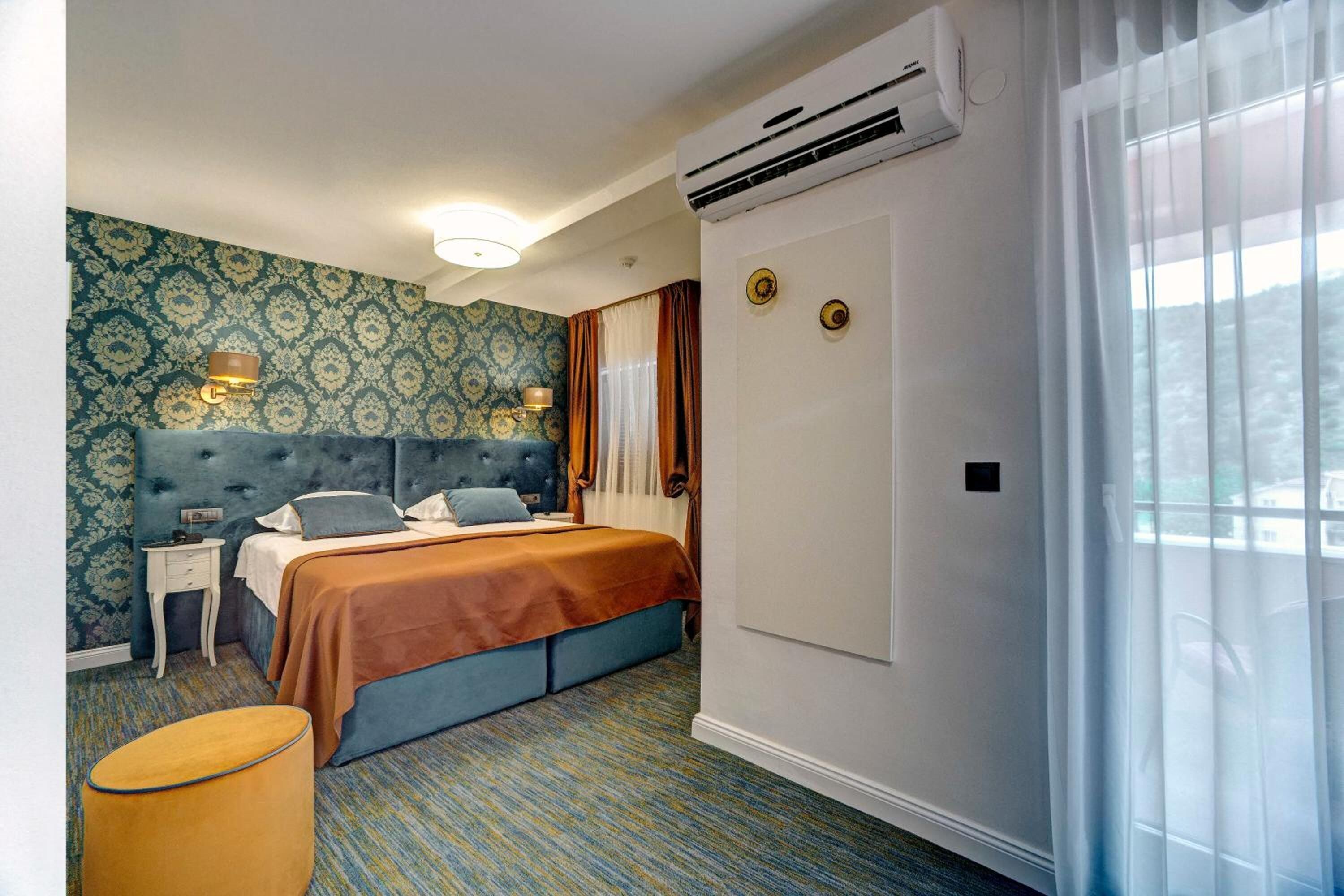 Habitació d'hotel amb un llit, una tauleta de nit, un aparell d'aire condicionat, una finestra amb cortines i un paper pintat amb un disseny.