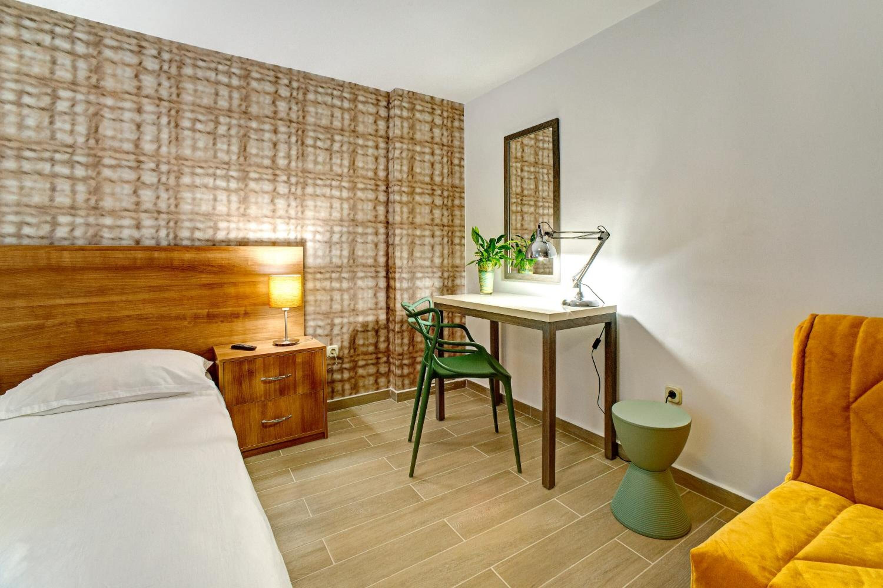 Habitación de hotel con cama, escritorio con lámpara, silla, mesita y taburete redondo.