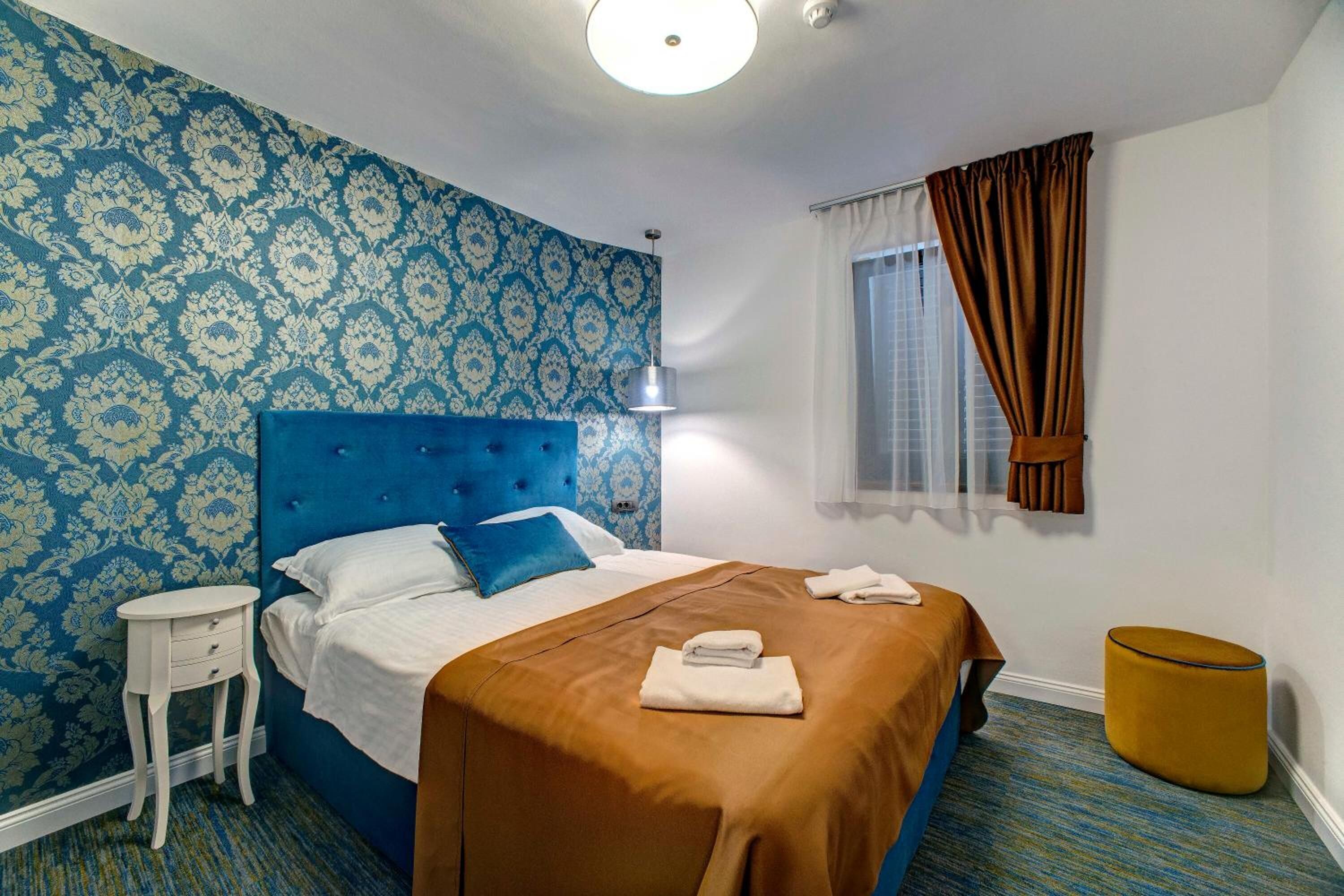 A hotel room with a blue patterned wallpaper, a tufted headboard, a single bed with white and blue bedding, a small white nightstand, a round yellow ottoman, and a window with brown curtains.