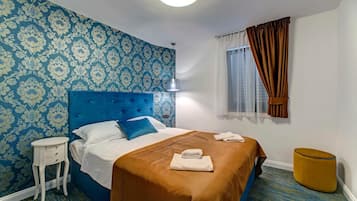 A hotel room with a blue patterned wallpaper, a tufted headboard, a single bed with white and blue bedding, a small white nightstand, a round yellow ottoman, and a window with brown curtains.