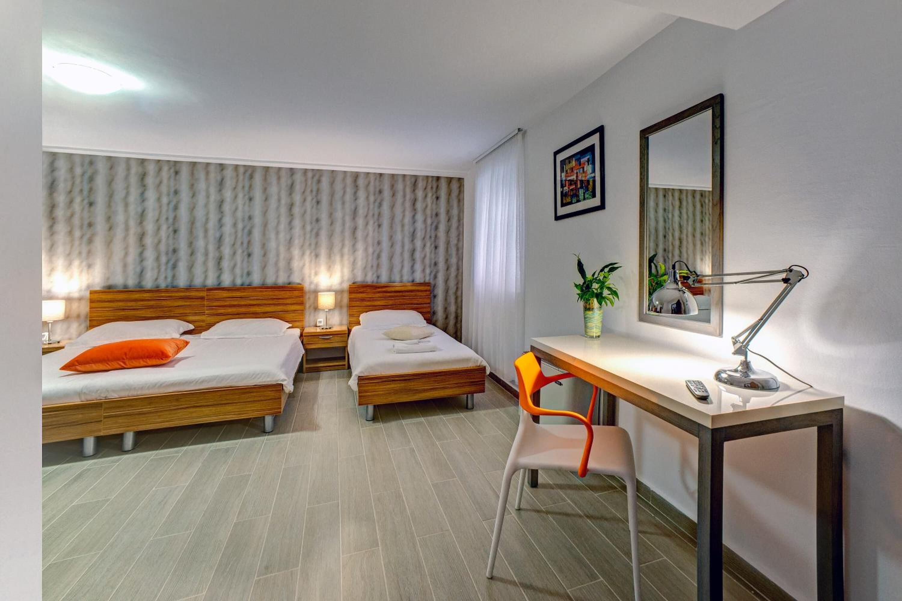 A hotel room with two beds, a desk, a chair, a desk lamp, a mirror, and a potted plant.