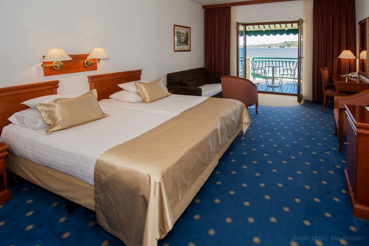 A hotel room with a large bed, a desk, a chair, and a balcony with a view.